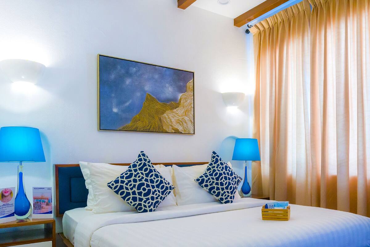 Una habitación de hotel con una cama, dos lámparas de noche, un cuadro y una ventana con cortinas.