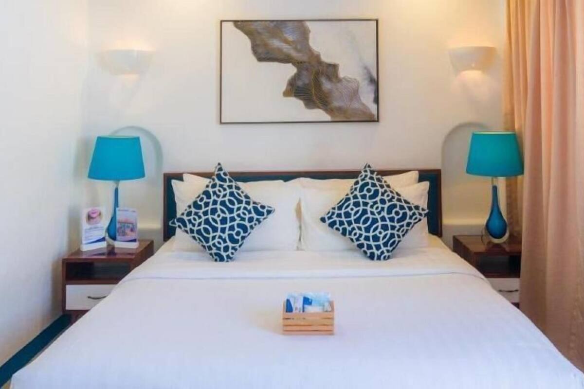 Habitación de hotel con cama, dos mesitas de noche, una obra de arte colgada en la pared y una mesita de madera con un recipiente de artículos de tocador.