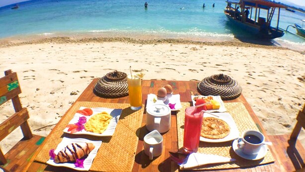 A beach table with breakfast items, a drink, and a view of the sea with boats. - La Bella - Hotel Villa & Spa (Gili Trawangan)