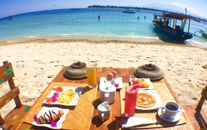 Une table installée sur la plage, avec des aliments pour le petit-déjeuner, une boisson, et une vue sur la mer et les bateaux.
