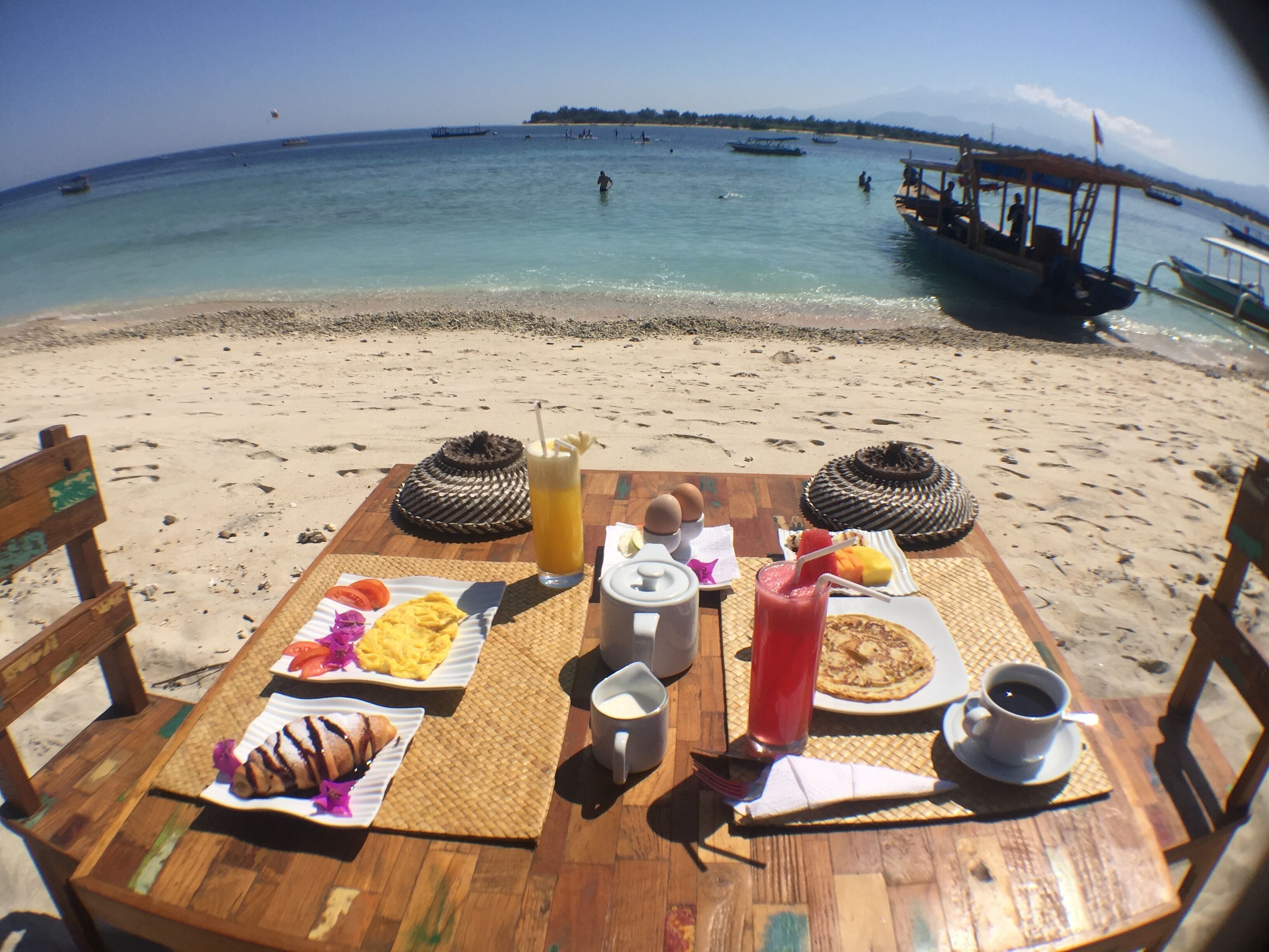 Een houten tafel met ontbijtspullen en uitzicht op het strand en boten.