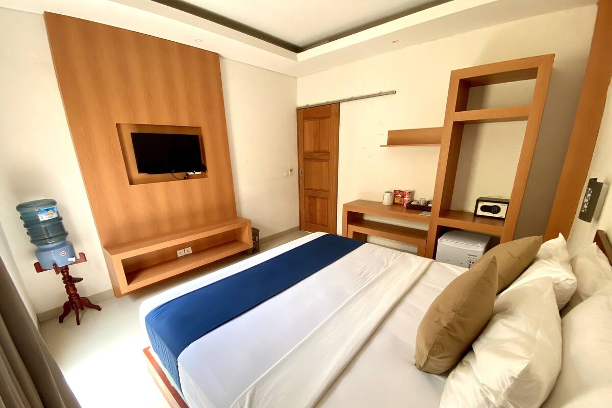 Ein Schlafzimmer mit einem Bett, einem an der Wand befestigten Fernseher, einem Holzschrank, einem Schreibtisch mit Stuhl und einem Wasserspender.