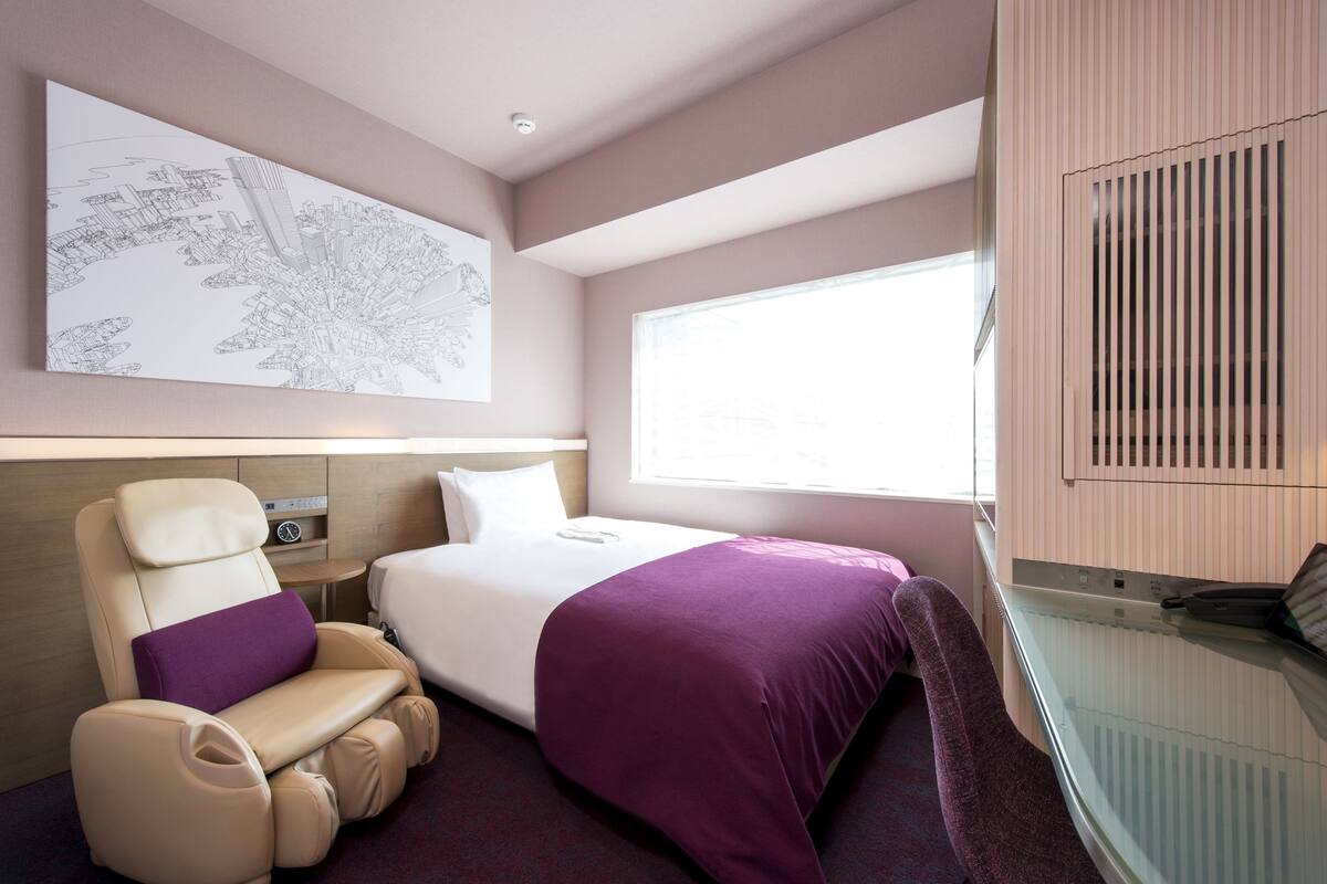 Habitación de hotel con cama, sillón de masajes, un cuadro con motivo de paisaje urbano y un ventanal grande.