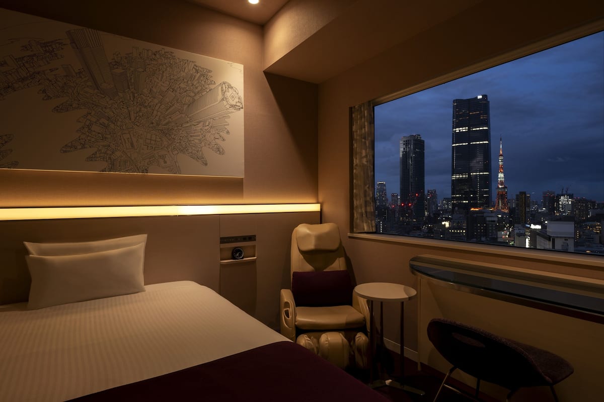 Habitación de hotel con cama, una silla, una mesita y vistas a un paisaje urbano nocturno.