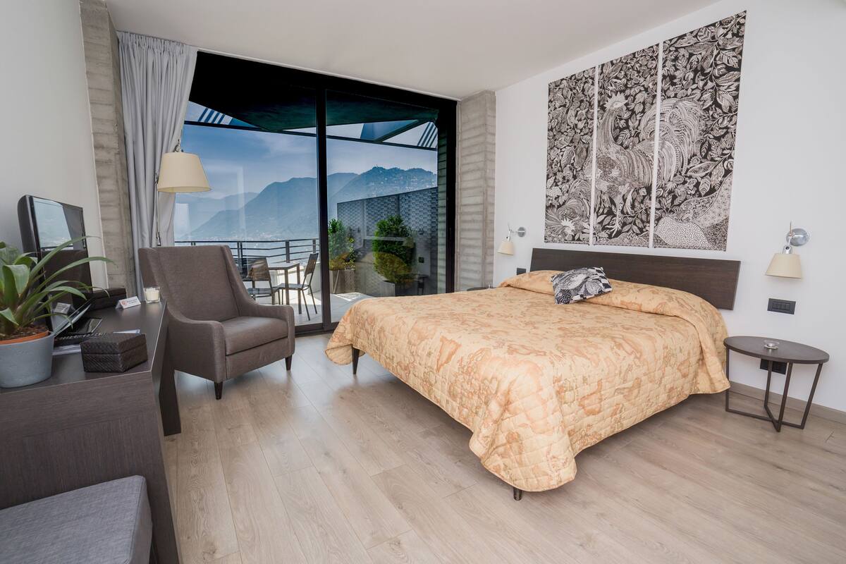 A modern hotel room with a large bed, a desk with a laptop, a chair, a small table, and a potted plant.