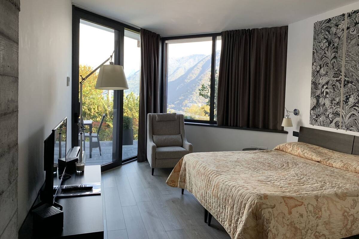 A hotel room with a bed, a desk with a computer, a chair, a TV, and a view of mountains through large windows.