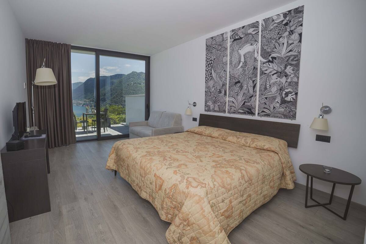A hotel room with a bed, a desk, a chair, a small table, and a large window with a view of mountains.
