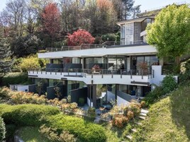 Una casa moderna de dos pisos con balcón, rodeada de abundante follaje y árboles.