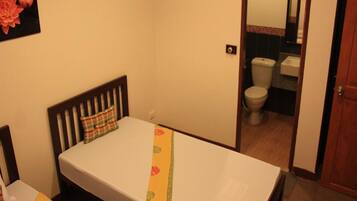 A small, single-bed room with a wooden headboard, a bedside table, and a plain bedspread. A bathroom is visible through an open door, featuring a toilet and a sink.
