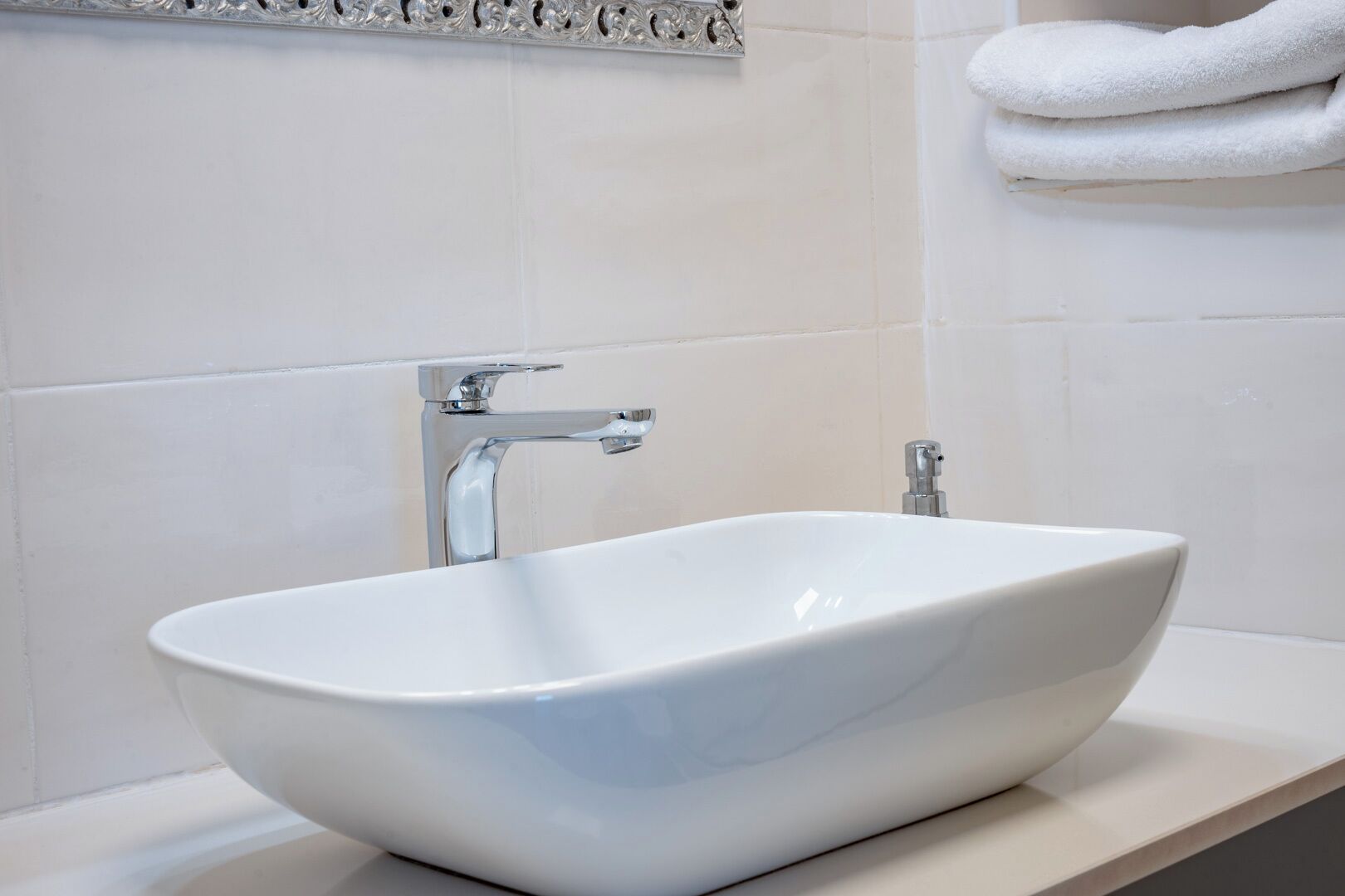 A white vessel sink with a chrome faucet in a bathroom with white tiles and a towel rack.