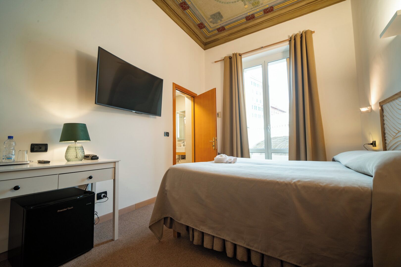 Habitación de hotel con cama, escritorio con lámpara, televisor y vistas al exterior.