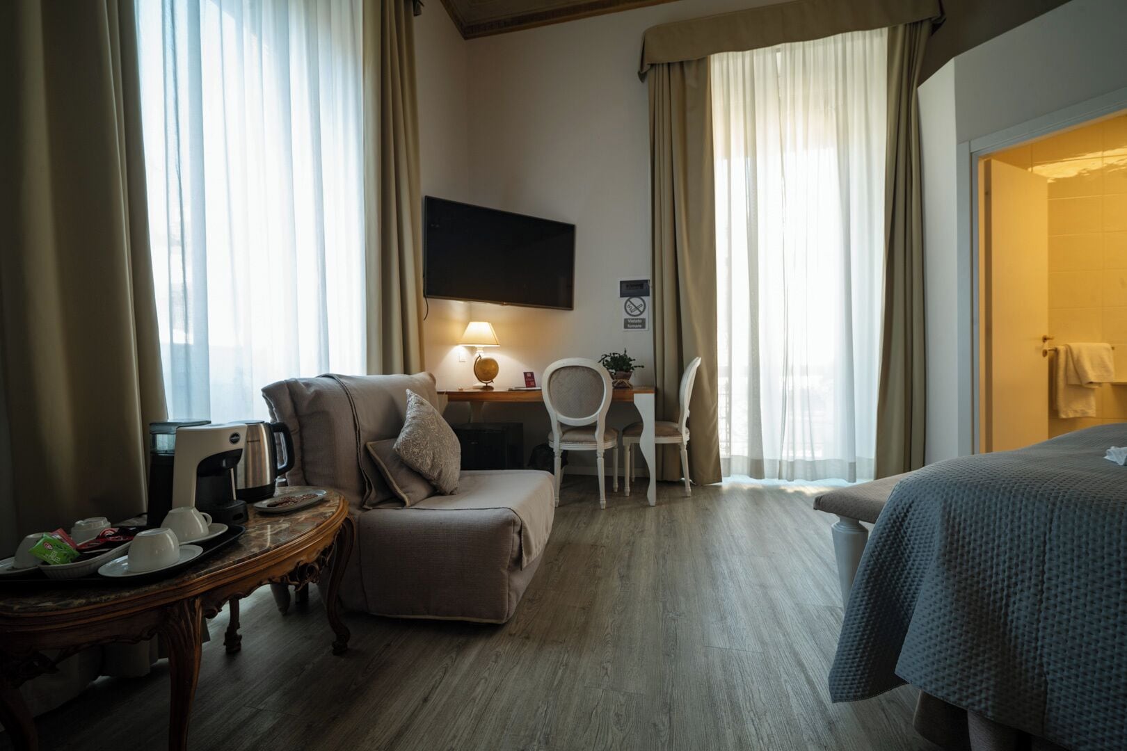 A hotel room with a bed, a desk with a lamp, a chair, a small table with a coffee maker, and curtains.