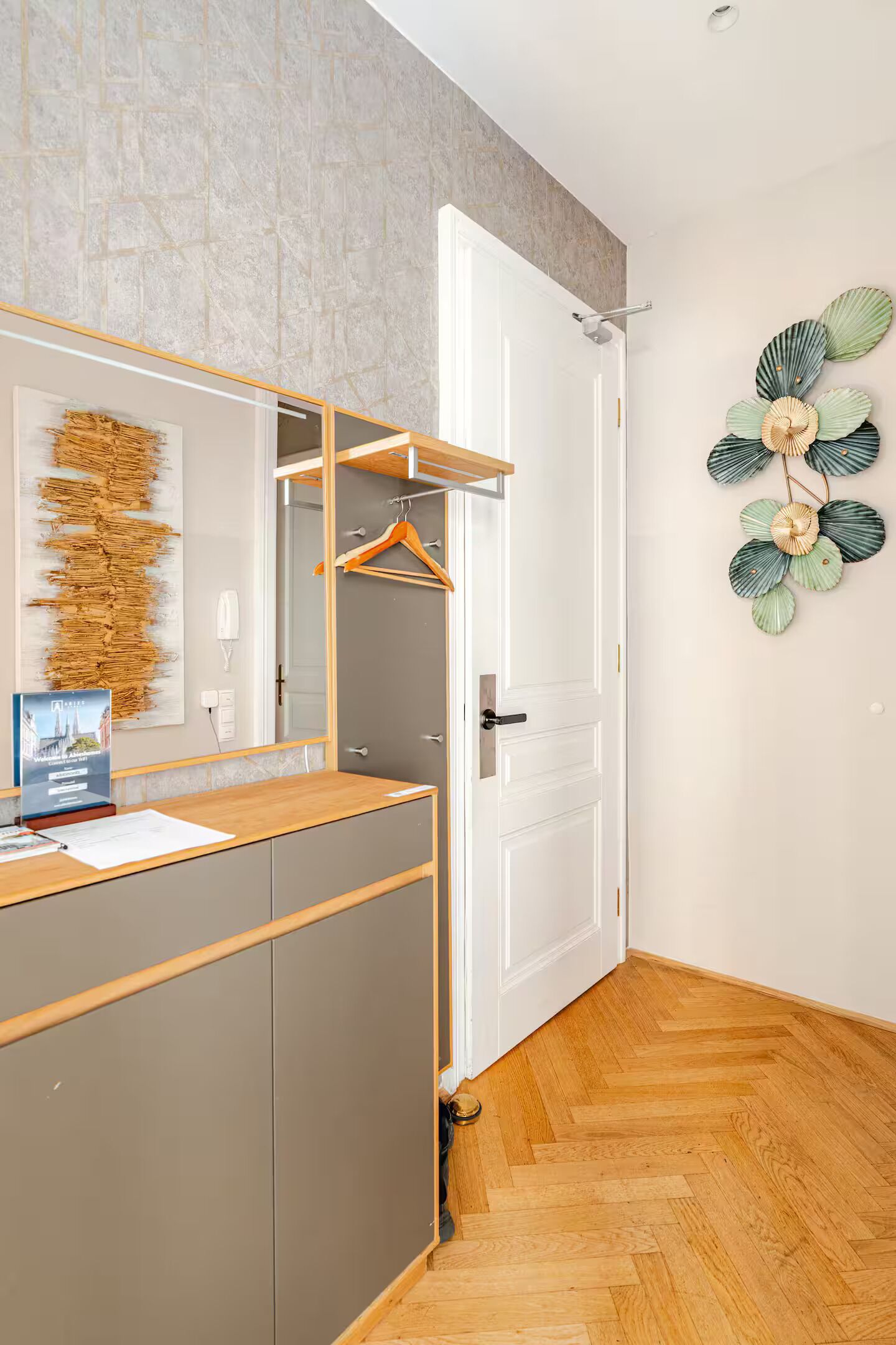 Habitación de hotel con piso de madera, un armario gris, un escritorio de madera con espejo y un ventilador decorativo fijado a la pared.