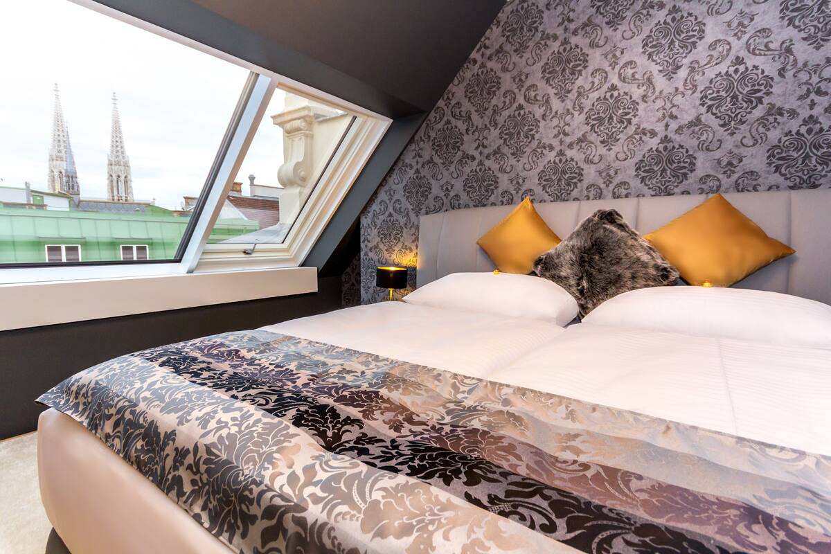 Une chambre avec un grand lit, une literie à motifs, une table de chevet avec une lampe et une fenêtre donnant sur des immeubles.