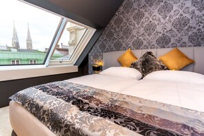 Un dormitorio con una cama grande, ropa de cama estampada, una mesita de noche con lámpara y una ventana con vistas a edificios.