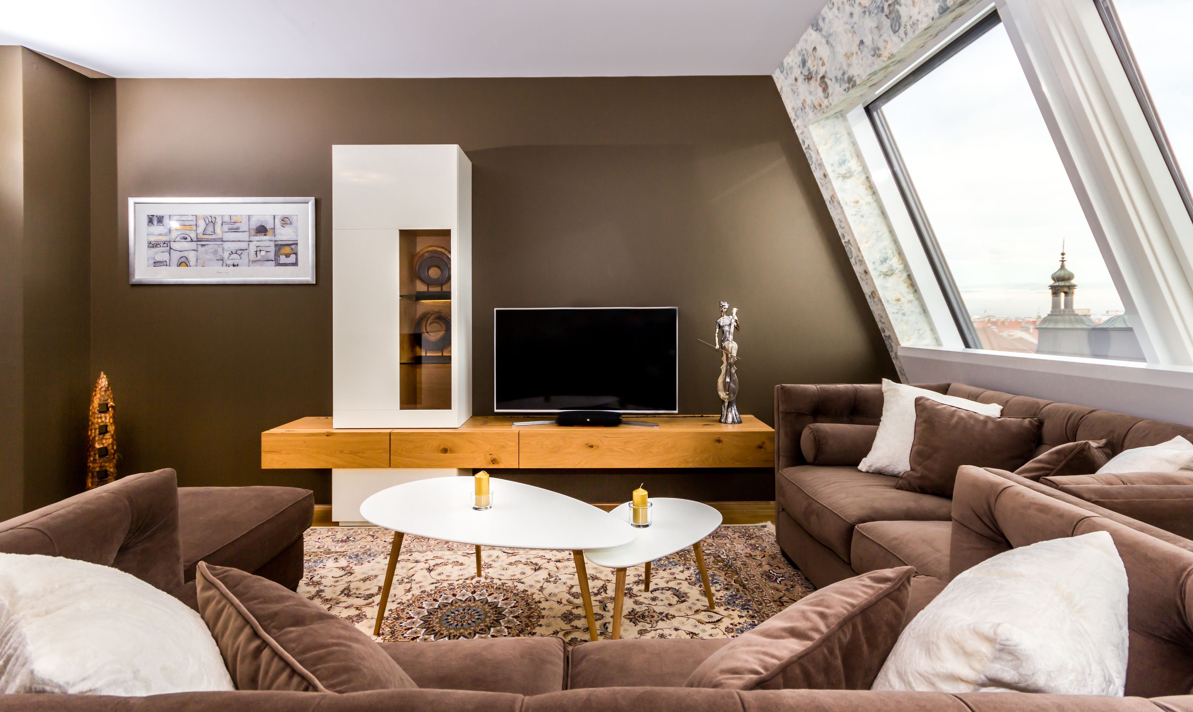 Een moderne woonkamer met een flatscreen televisie, een wit kastje, een zithoek en uitzicht op een stadsgezicht door een groot raam.