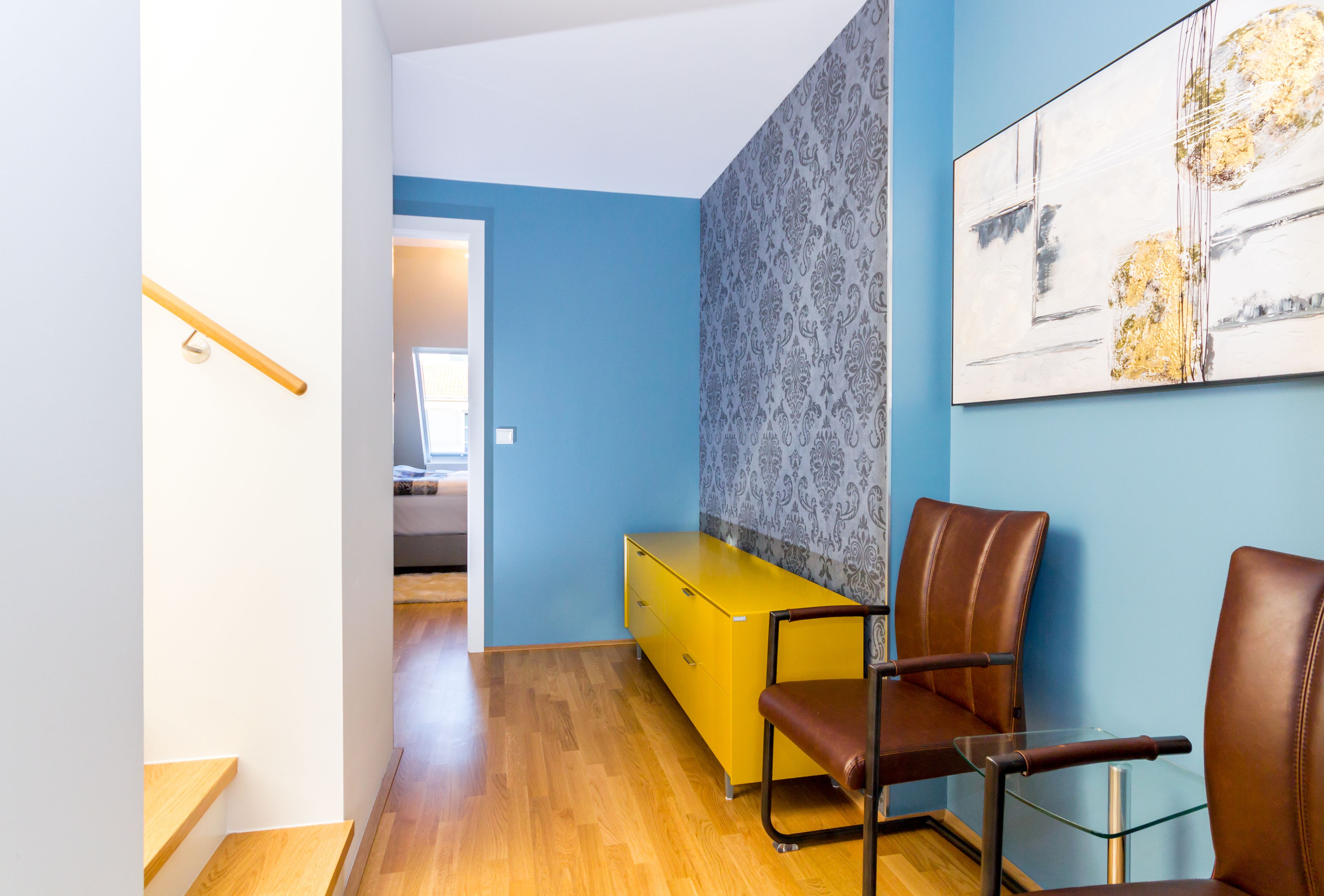 Een moderne kamer met een houten vloer, een gele ladebank, twee leren fauteuils en een groot abstract schilderij aan de muur.