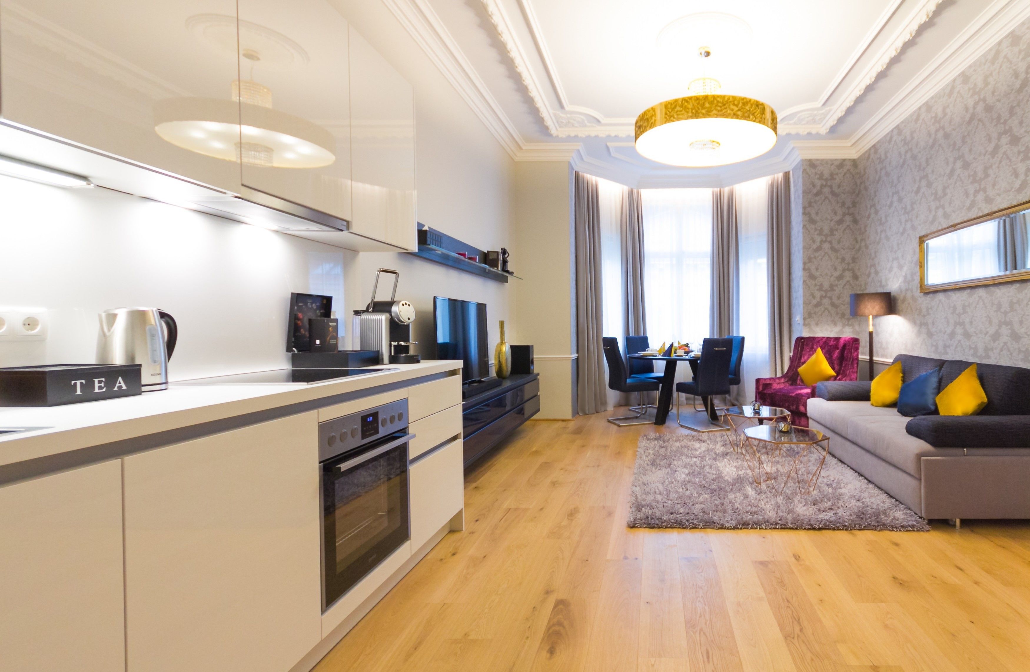 A modern kitchen with a built-in oven and a coffee machine, a dining area with a table and chairs, and a living area with a sofa and a lamp.