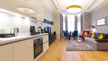 A modern kitchen with a built-in oven and a coffee machine, a dining area with a table and chairs, and a living area with a sofa and a lamp.