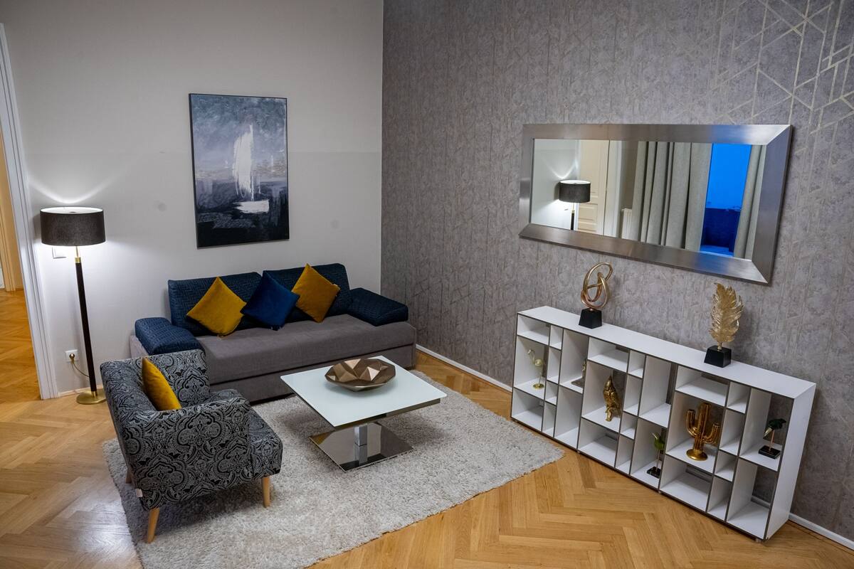 Un salon moderne avec un canapé gris, un fauteuil à motifs et une table basse blanche. On peut également apercevoir un grand miroir et un meuble console blanc agrémenté d’objets décoratifs.
