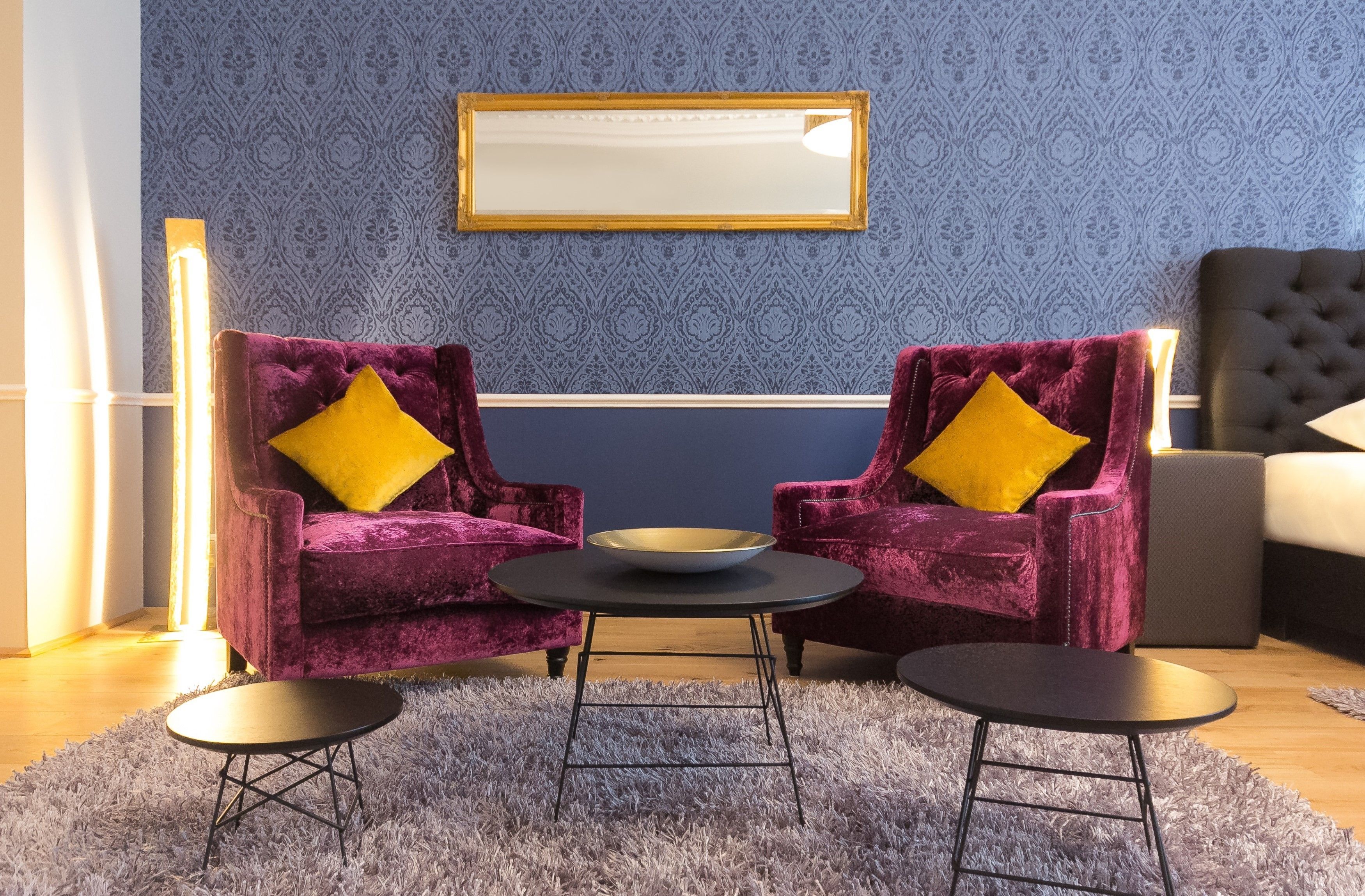 Una habitación con dos sillones morados de terciopelo, cojines amarillos, una mesita redonda y un espejo en la pared.