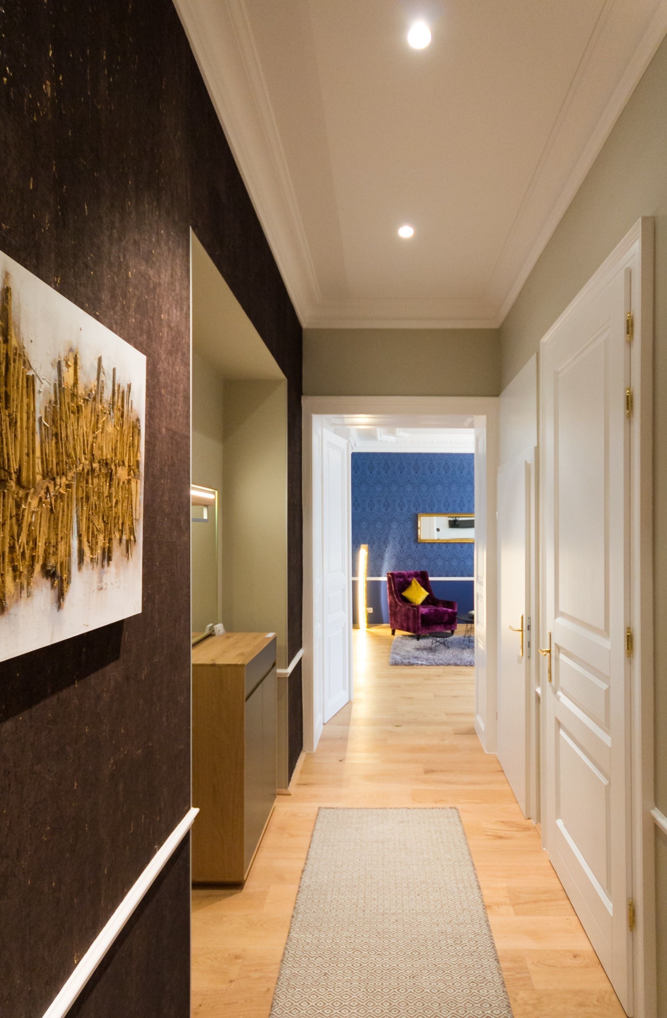 Un pasillo con piso de madera, un tapete gris, una pared oscura con una obra de arte texturizada y una puerta que da a una habitación con pared azul y una silla morada.