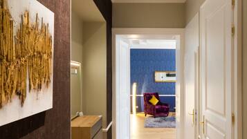 Un pasillo con piso de madera, un tapete gris, una pared oscura con una obra de arte texturizada y una puerta que da a una habitación con pared azul y una silla morada.
