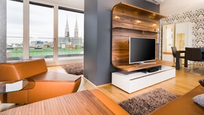 Een moderne woonkamer met een grote flatscreen televisie aan een houten wandpaneel, een eethoek met een tafel en stoelen, en uitzicht op een stadsbeeld met een kerktoren.
