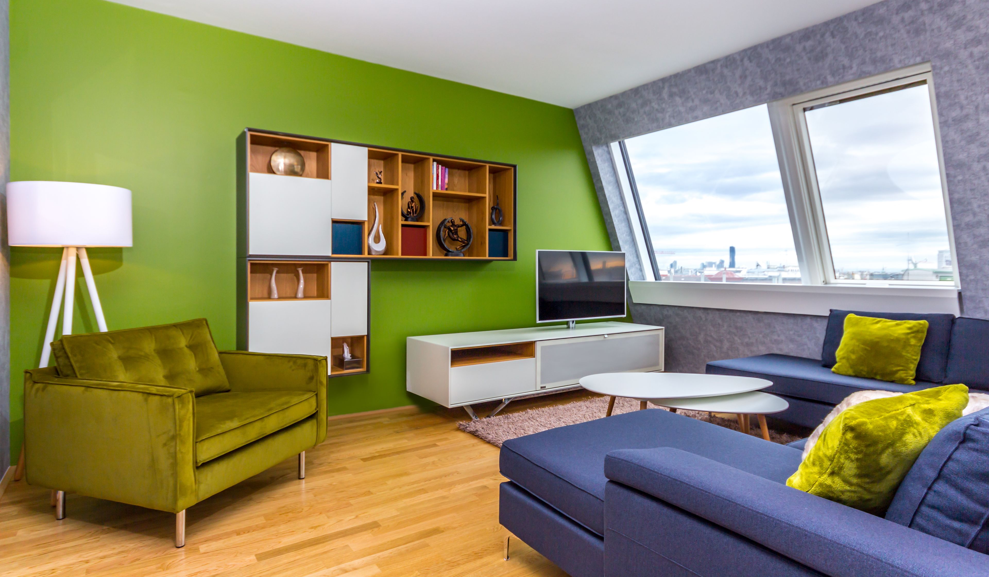 Una sala de estar moderna con una pared verde, un mueble blanco para entretenimiento, un sofá azul con cojines amarillos y vistas a través de un ventanal amplio.