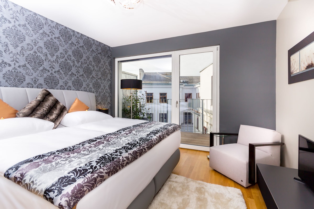 Quarto moderno com uma cama grande, uma cadeira, uma escrivaninha e varanda com vista para edifícios.