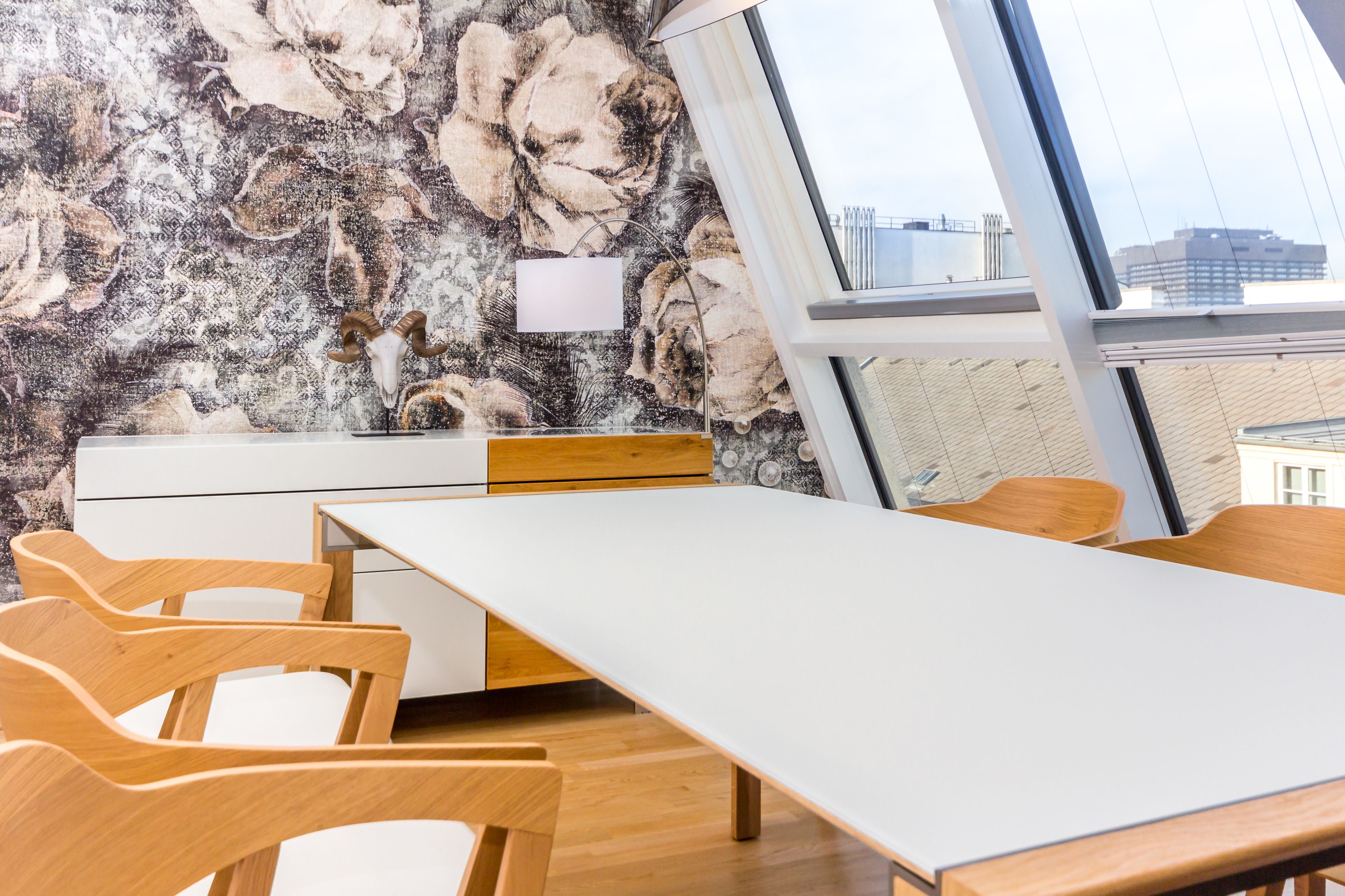 Uma sala de reunião moderna com uma mesa branca, cadeiras de madeira, um armário branco e uma grande janela com vista para prédios.