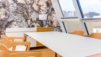 Uma sala de reunião moderna com uma mesa branca, cadeiras de madeira, um armário branco e uma grande janela com vista para prédios.