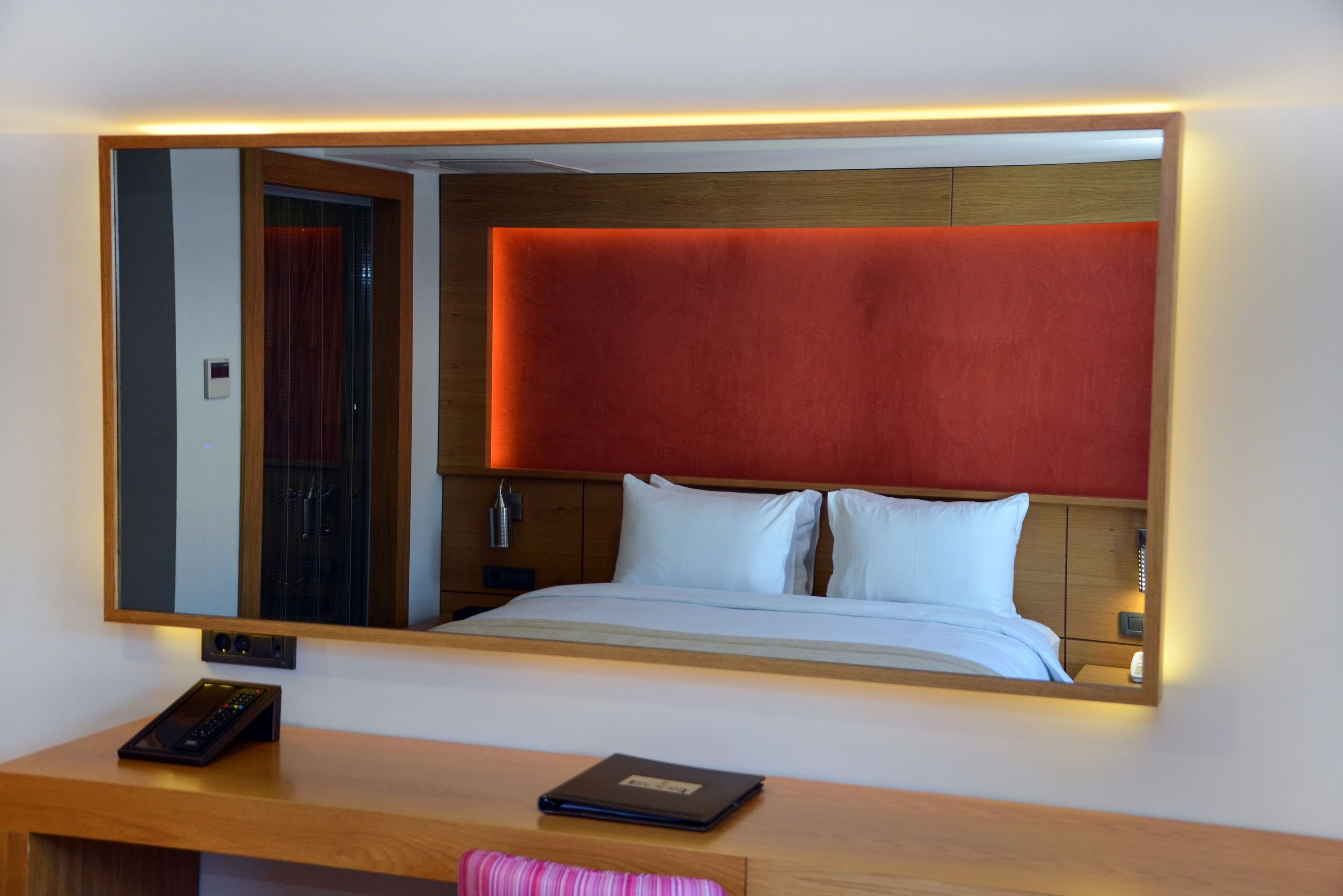 A hotel room with a large bed, a bedside table with a remote control and a book, and a mirror reflecting the room's interior.