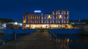 A waterfront hotel with a prominent sign, a wooden boardwalk leading to the entrance, and a calm river in the foreground. - Bacacan Otel (Ayvalik)