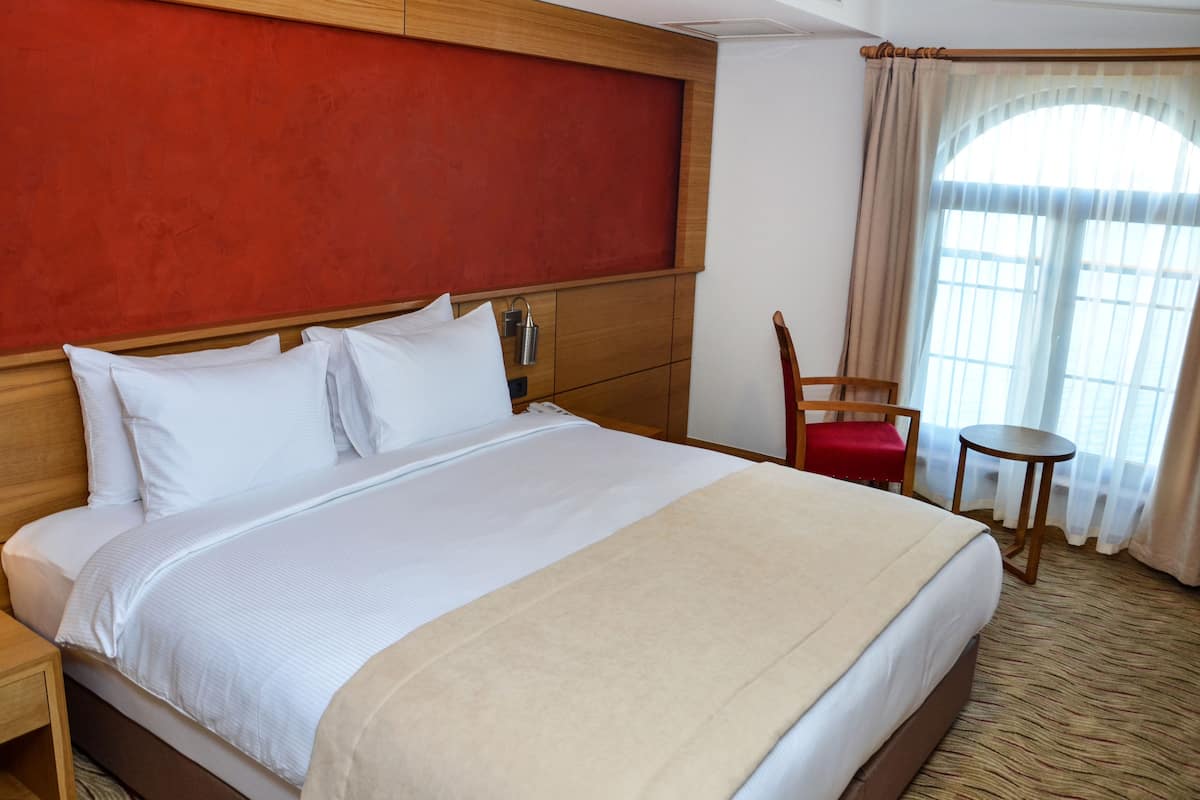 A hotel room with a large bed, a wooden headboard, a red chair, a small table, and a window with curtains.