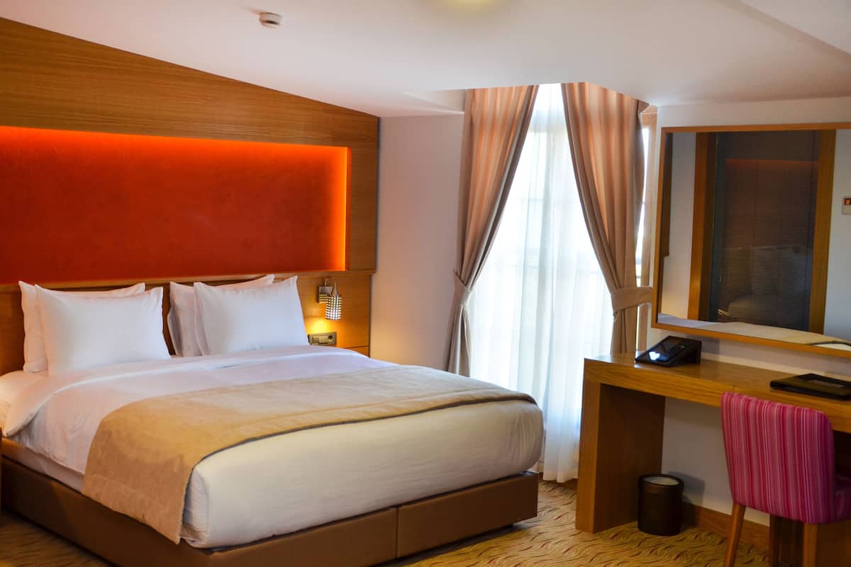 A hotel room with a large bed, a wooden desk, a chair, a mirror, and a window with curtains.