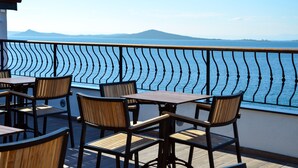 A terrace with wooden tables and chairs, a decorative railing, and a view of the sea and distant mountains. - Bacacan Otel (Ayvalik)