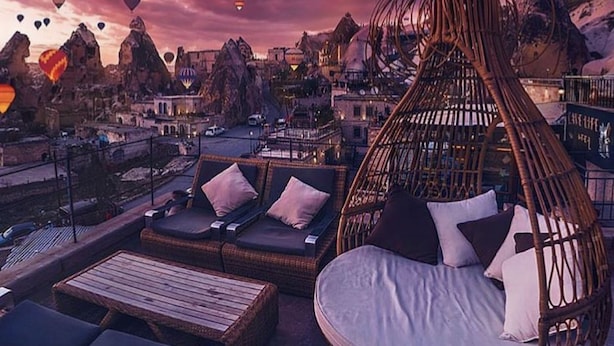 A rooftop lounge with wicker furniture, a view of a cityscape, and hot air balloons in the sky. - The Owl Cave Hotel (Nevsehir)