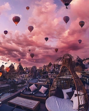 Un salón en la azotea con mobiliario de mimbre, vista panorámica de la ciudad y globos aerostáticos en el cielo.