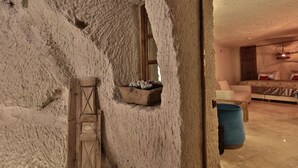 A room with a textured stone wall, a wooden ladder, a window, a bed with a patterned bedspread, a chair, and a blue barrel. - The Owl Cave Hotel (Nevsehir)