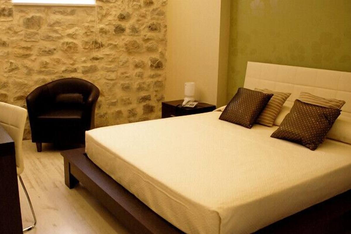 Ein Schlafzimmer mit einer Steinwand, einem Bett mit vielen Kissen, einem Sessel, einem Nachttisch und einer Vase mit Blumen.