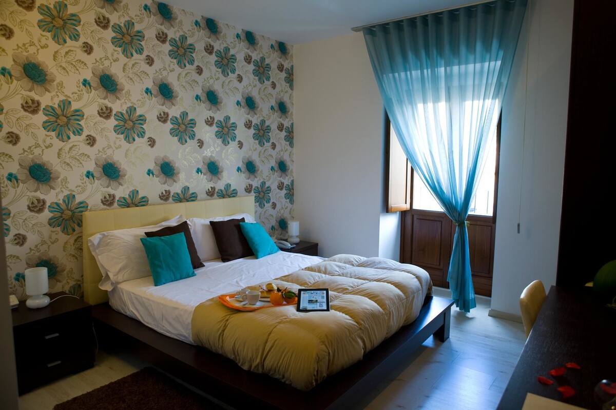 Ein Schlafzimmer mit einem großen Bett, floralem Wandpapier, einem Nachttisch mit Lampe und einem Fenster mit blauen Vorhängen.