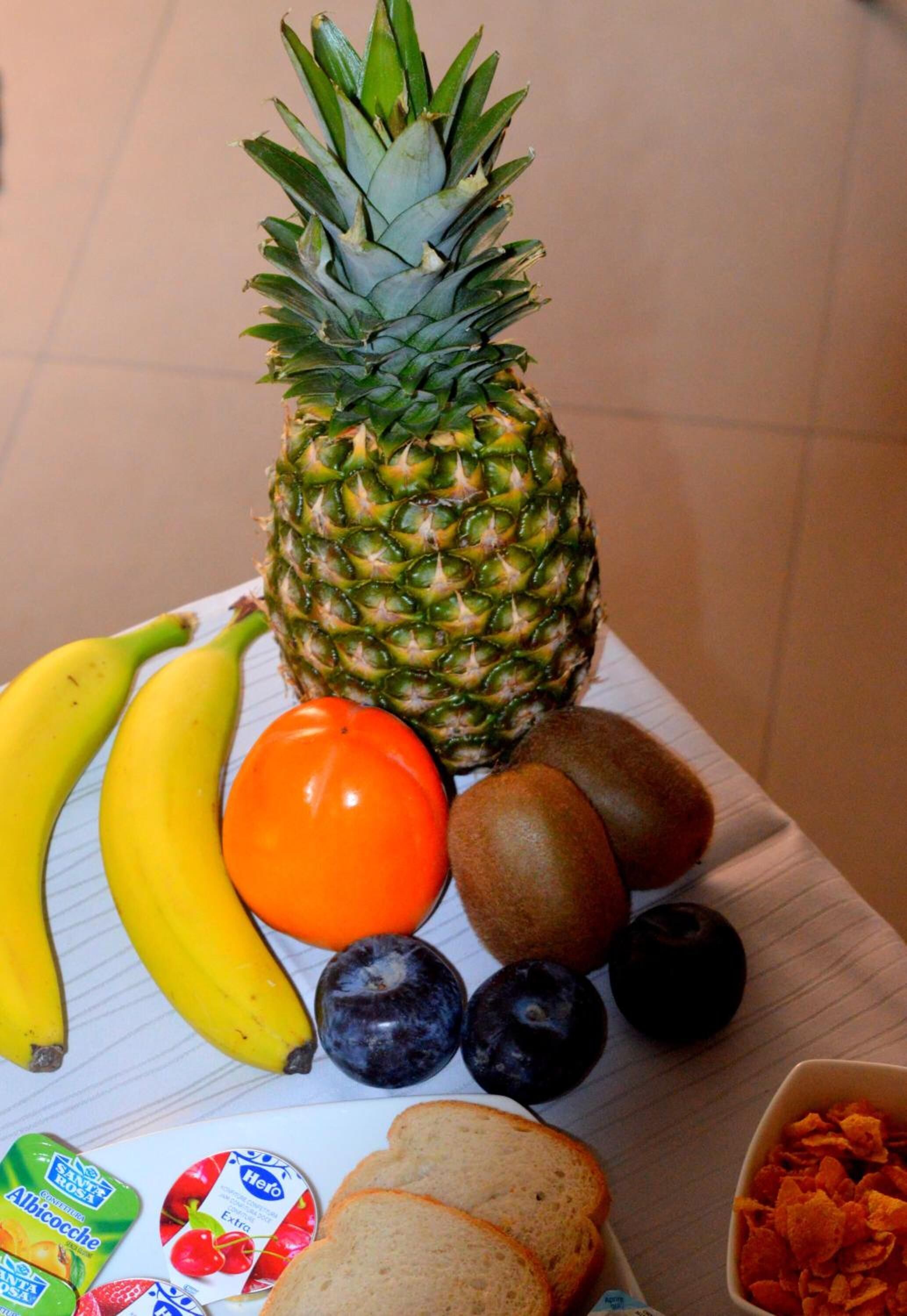 Ett bord med ett urval av frukter och snacks, inklusive ananas, bananer, en tomat, kiwifrukter, blåbär och flingor.