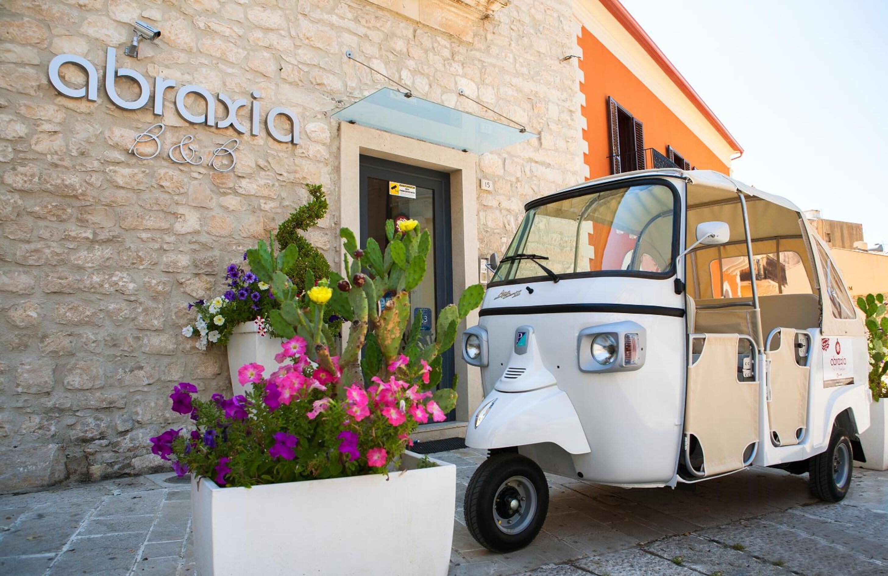 Un véhicule blanc à trois roues est garé devant un immeuble à la façade en pierre, sur lequel on peut lire une enseigne indiquant « Abraxia, chambres d’hôtes ».