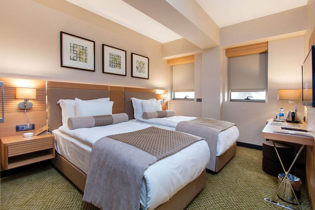 A hotel room with two beds, a desk, a chair, a lamp, and framed artwork on the wall.