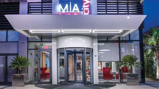 Une entrée d’hôtel moderne avec une porte tournante, une façade en verre et des sièges rouges à l’extérieur.