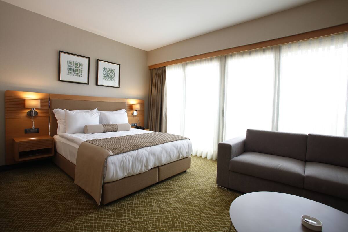A hotel room with a large bed, a sofa, and a small table. There are two framed pictures on the wall, a nightstand with a lamp, and a window with curtains.