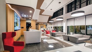 Un hall d'entrée moderne avec un comptoir d'accueil, des fauteuils rouges, des canapés gris et une grande fenêtre donnant sur l'extérieur.