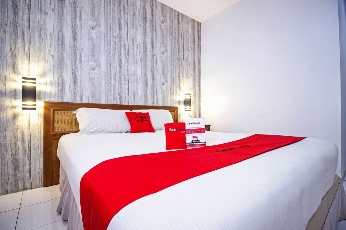 A hotel room with a bed, a wooden headboard, a red throw blanket, and two wall-mounted light fixtures.