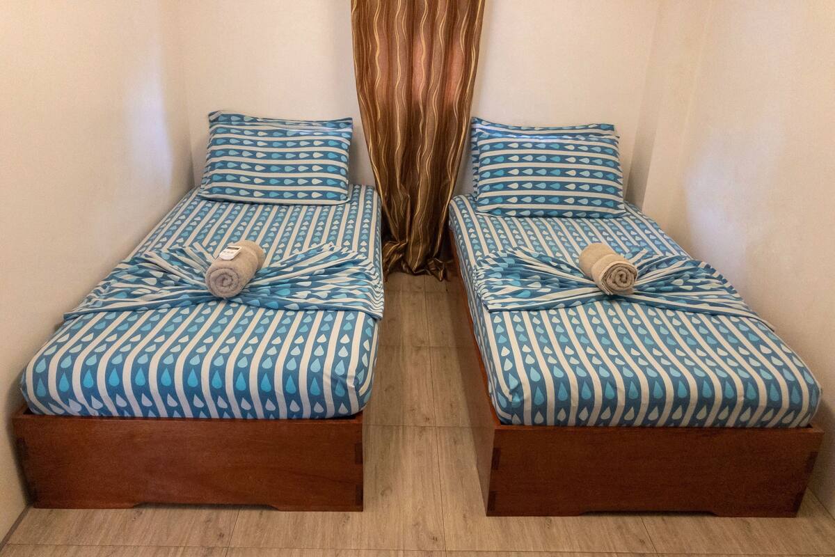 Two single beds with matching blue and white patterned bedding and wooden bases, positioned side by side with a curtain in the background.